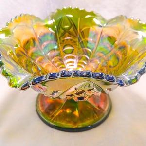 May include: Iridescent glass pedestal bowl with a ruffled edge. The bowl features a vibrant display of colors, including green, yellow, and orange, with a decorative pattern. The base is a matching iridescent color. A decorative piece for display.