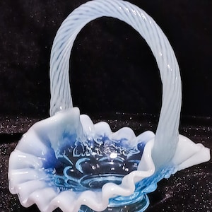 Op de afbeelding: Een lichtblauwe glazen mand met een gedraaide handgreep en gegolfde randen. De mand heeft een ronde basis en een gedetailleerd bloemmotief aan de binnenkant. Het glas heeft een mat uiterlijk met een verloop van blauw en wit.