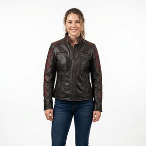 May include: A dark brown leather jacket featuring a stand-up collar, a full-length front zip, and two zipped chest pockets. The sleeves and shoulders are accented with intricate red embroidery patterns. The jacket is paired with blue denim jeans.