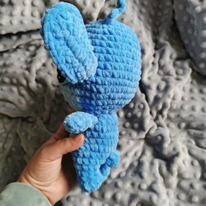 Puede incluir: Un juguete de conejo de ganchillo hecho a mano, de color azul claro. El conejo de peluche tiene orejas grandes, brazos y piernas peque&ntilde;os y una cola rizada. El juguete est&aacute; hecho de hilo suave y texturizado y tiene ojos negros. El conejo se sostiene en una mano.