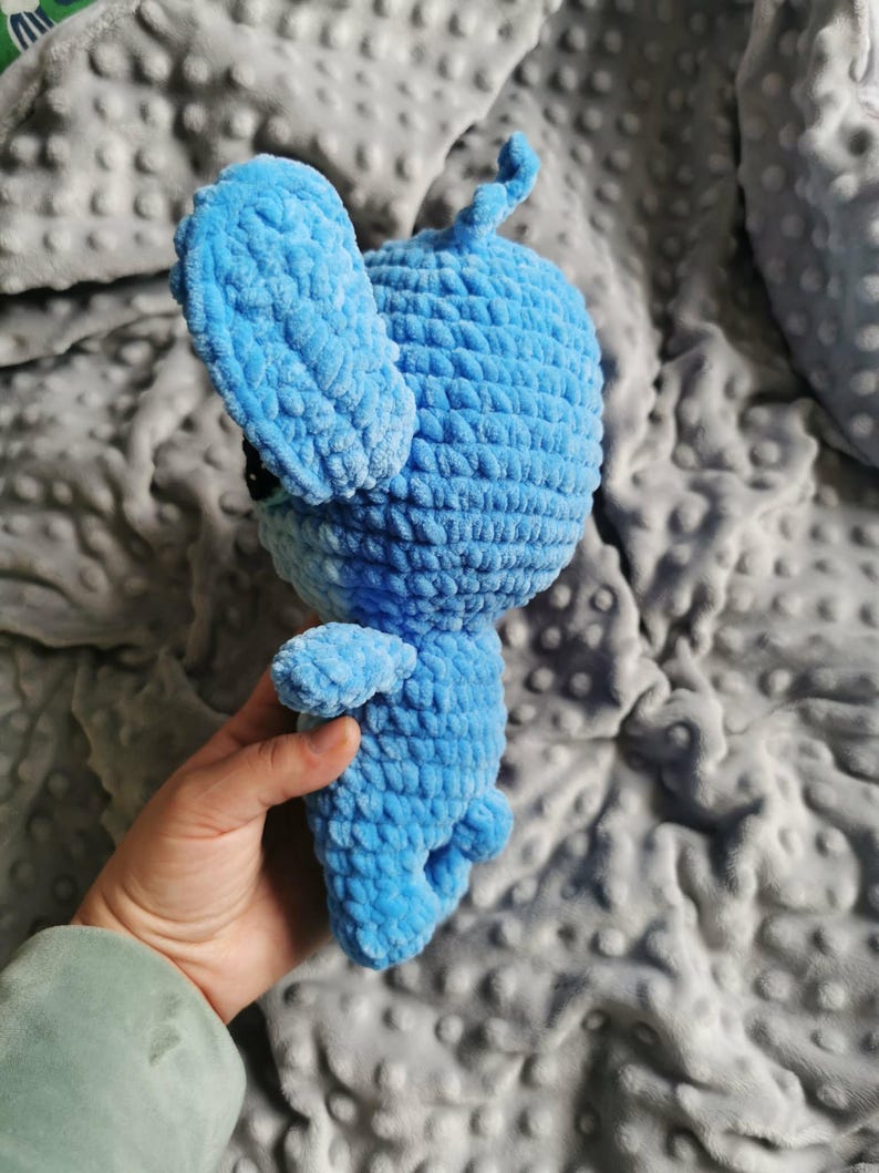 Puede incluir: Un juguete de conejo de ganchillo hecho a mano, de color azul claro. El conejo de peluche tiene orejas grandes, brazos y piernas peque&ntilde;os y una cola rizada. El juguete est&aacute; hecho de hilo suave y texturizado y tiene ojos negros. El conejo se sostiene en una mano.