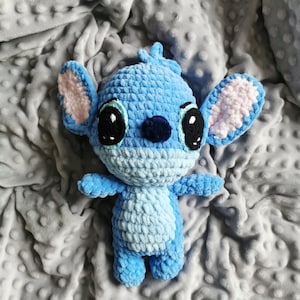 Puede incluir: Una mu&ntilde;eca Stitch azul hecha a mano con ganchillo, con ojos negros grandes, orejas internas rosas y una nariz azul oscuro. El peluche est&aacute; sobre una manta gris texturizada. La mu&ntilde;eca tiene brazos y piernas peque&ntilde;os.