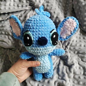 Puede incluir: Un peluche azul hecho a mano con ganchillo, con grandes ojos negros, interior de las orejas rosa y una nariz azul oscuro. El juguete tiene un aspecto texturizado y difuso, y se sostiene sobre un fondo gris.