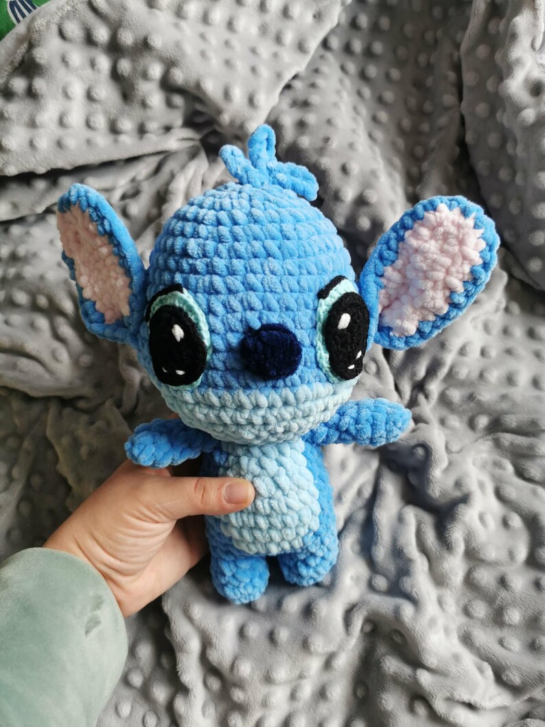 Puede incluir: Un peluche azul hecho a mano con ganchillo, con grandes ojos negros, interior de las orejas rosa y una nariz azul oscuro. El juguete tiene un aspecto texturizado y difuso, y se sostiene sobre un fondo gris.