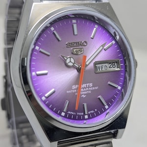 May include: A Seiko 5 Sports automatic wristwatch with a metallic purple sunburst dial, silver-tone hour markers, and a bright orange second hand. The watch features a day-date window displaying WED 28, a stainless steel case, and a matching link bracelet.