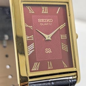 Könnte beinhalten: Eine rechteckige Seiko Quartz Armbanduhr mit goldfarbenem Gehäuse und einem tiefroten Zifferblatt mit texturiertem Wellenmuster. Das Zifferblatt zeigt goldene römische Ziffern, goldene Zeiger und das Seiko Quartz Logo. Mit schwarzem Lederarmband.