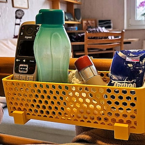 Könnte beinhalten: Ein gelber Nachttisch-Organizer aus Kunststoff mit Wabenmuster, der eine grüne Wasserflasche, ein Telefon, eine Tube und eine blaue Taschentuchbox enthält. Der Organizer ist an einem hölzernen Bettrahmen befestigt.