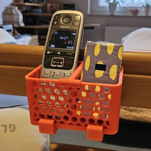 Könnte beinhalten: Ein orangefarbener, 3D-gedruckter Nachttisch-Organizer mit Wabenmuster, der ein silbernes und schwarzes Gigaset-Telefon und eine lila Schachtel mit gelben Ovalen enthält. Der Organizer ist an einem hölzernen Bettrahmen befestigt.