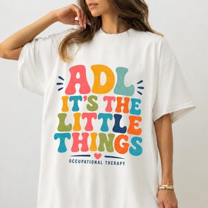 May include: A white short-sleeve t-shirt featuring a multi-colored graphic print with the text ADL IT'S THE LITTLE THINGS and OCCUPATIONAL THERAPY. The letters are in a bold, rounded font with various colors including red, yellow, blue, and green.
