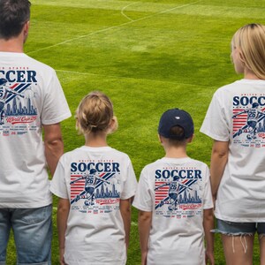 Op de afbeelding: Witte t-shirts met een afbeelding van een voetballer, de tekst United States Soccer World Cup 26, en een lijst met gaststeden waaronder New York, New Jersey, Philadelphia en Seattle, voorzien van rode en blauwe strepen.