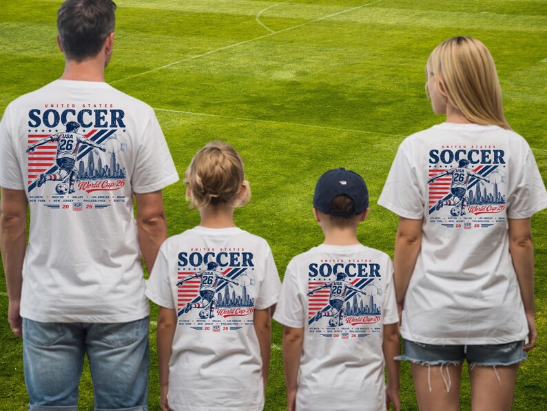Op de afbeelding: Witte t-shirts met een afbeelding van een voetballer, de tekst United States Soccer World Cup 26, en een lijst met gaststeden waaronder New York, New Jersey, Philadelphia en Seattle, voorzien van rode en blauwe strepen.