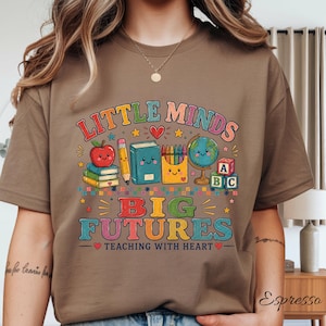 Peut inclure: Un t-shirt marron avec un motif color&eacute; portant le texte "LITTLE MINDS BIG FUTURES TEACHING WITH HEART" et des illustrations d'une pomme souriante, de livres, d'un crayon, de crayons de couleur, d'un globe et de cubes alphabet, avec des &eacute;toiles.