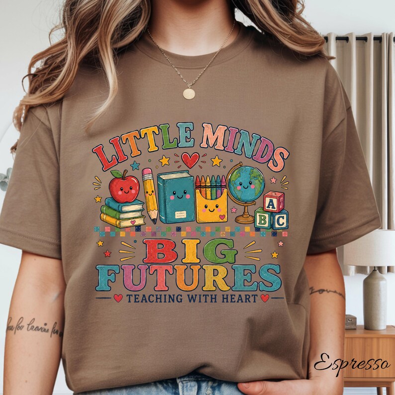 Peut inclure: Un t-shirt marron avec un motif color&eacute; portant le texte "LITTLE MINDS BIG FUTURES TEACHING WITH HEART" et des illustrations d'une pomme souriante, de livres, d'un crayon, de crayons de couleur, d'un globe et de cubes alphabet, avec des &eacute;toiles.