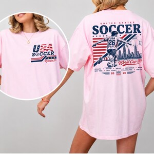Op de afbeelding: Een lichtroze t-shirt met een USA Soccer World Cup 26 afbeelding. De borstzijde toont een klein logo, terwijl de achterkant een grote illustratie van een voetballer, een skyline, de tekst United States Soccer en diverse stadsnamen bevat.