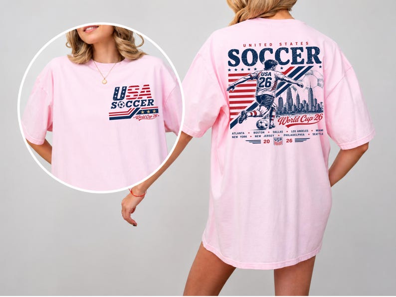 Op de afbeelding: Een lichtroze t-shirt met een USA Soccer World Cup 26 afbeelding. De borstzijde toont een klein logo, terwijl de achterkant een grote illustratie van een voetballer, een skyline, de tekst United States Soccer en diverse stadsnamen bevat.