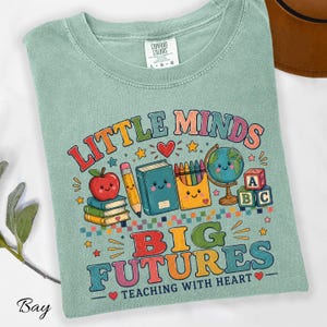 Peut inclure: Un t-shirt vert sauge avec un motif color&eacute; portant le texte "LITTLE MINDS BIG FUTURES TEACHING WITH HEART". Le design comprend des illustrations d'une pomme souriante, d'un crayon, d'un livre, d'une bo&icirc;te de crayons, d'un globe et de cubes alphabet.