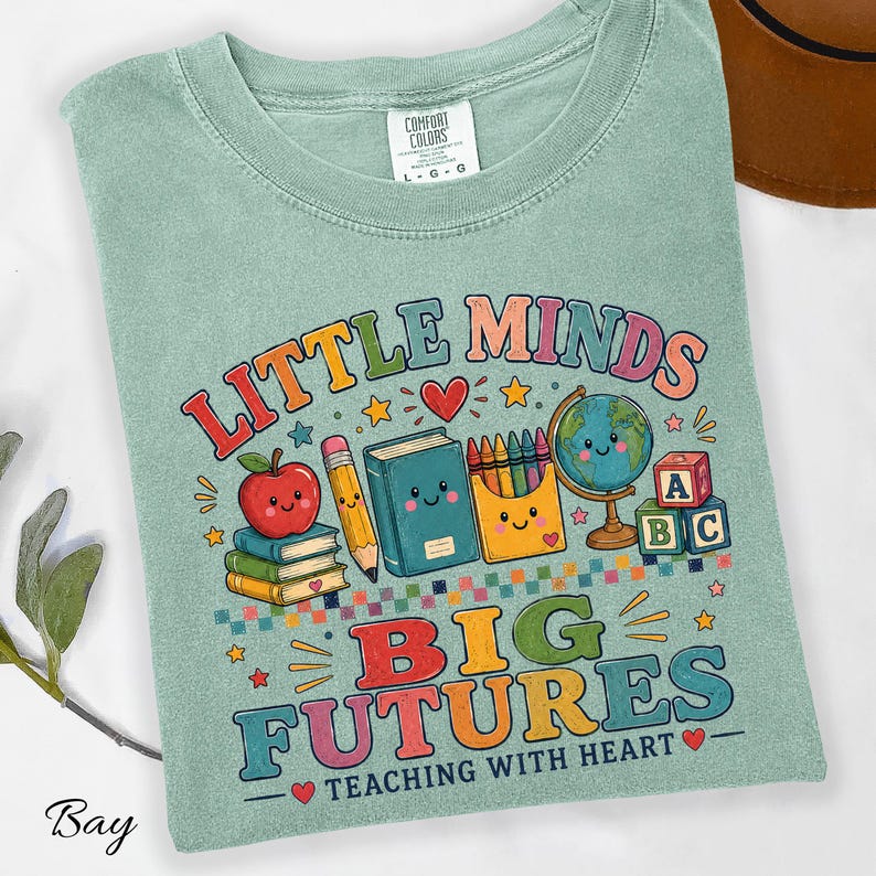 Peut inclure: Un t-shirt vert sauge avec un motif color&eacute; portant le texte "LITTLE MINDS BIG FUTURES TEACHING WITH HEART". Le design comprend des illustrations d'une pomme souriante, d'un crayon, d'un livre, d'une bo&icirc;te de crayons, d'un globe et de cubes alphabet.