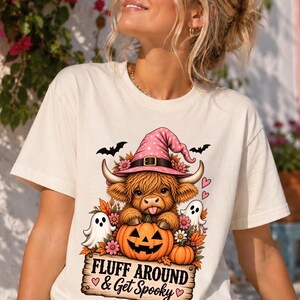 May include: A cream-colored t-shirt featuring a graphic of a Highland cow wearing a pink witch hat, sitting behind a jack-o'-lantern. The design includes two ghosts, autumn leaves, bats, and the text "FLUFF AROUND & Get Spooky" on a wooden sign.
