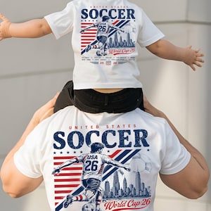 Op de afbeelding: Een wit t-shirt met een afbeelding van een voetballer, de tekst United States Soccer en World Cup 26. Het ontwerp bevat een rood-blauw gestreepte achtergrond, een stadssilhouet en een voetbal. Het t-shirt toont het nummer 26.
