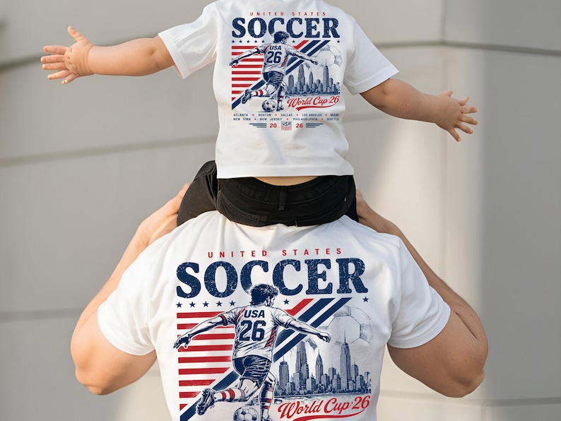 Op de afbeelding: Een wit t-shirt met een afbeelding van een voetballer, de tekst United States Soccer en World Cup 26. Het ontwerp bevat een rood-blauw gestreepte achtergrond, een stadssilhouet en een voetbal. Het t-shirt toont het nummer 26.