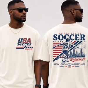 Op de afbeelding: Een off-white t-shirt met een klein USA Soccer World Cup 26 logo op de borst. De achterkant toont een grote afbeelding van een voetballer, een skyline, de tekst United States Soccer World Cup 26 en de namen van de gaststeden.