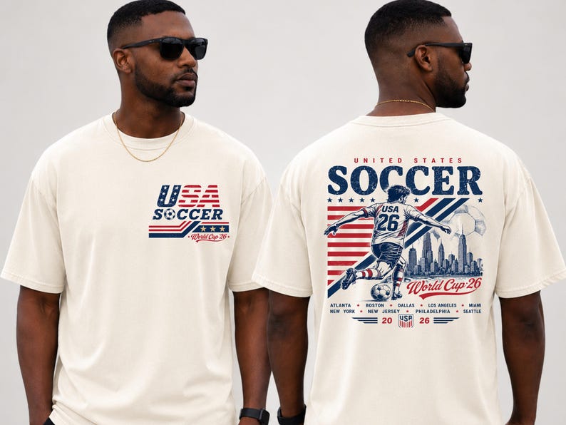 Op de afbeelding: Een off-white t-shirt met een klein USA Soccer World Cup 26 logo op de borst. De achterkant toont een grote afbeelding van een voetballer, een skyline, de tekst United States Soccer World Cup 26 en de namen van de gaststeden.