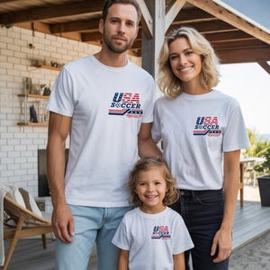 Op de afbeelding: Drie witte t-shirts met een afbeelding op de linkerborst. De afbeelding toont de tekst USA SOCCER in blauw en rood, met een klein voetbalicoontje en horizontale strepen. De t-shirts hebben een ronde hals en korte mouwen.