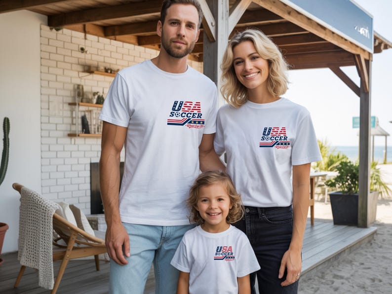 Op de afbeelding: Drie witte t-shirts met een afbeelding op de linkerborst. De afbeelding toont de tekst USA SOCCER in blauw en rood, met een klein voetbalicoontje en horizontale strepen. De t-shirts hebben een ronde hals en korte mouwen.