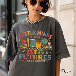 Peut inclure: Un t-shirt gris anthracite arborant un motif color&eacute; avec le texte "LITTLE MINDS BIG FUTURES TEACHING WITH HEART" et des illustrations d'une pomme souriante, de livres, d'un crayon, de crayons de couleur, d'un globe et de cubes alphabet.