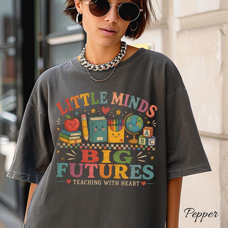 Peut inclure: Un t-shirt gris anthracite arborant un motif color&eacute; avec le texte "LITTLE MINDS BIG FUTURES TEACHING WITH HEART" et des illustrations d'une pomme souriante, de livres, d'un crayon, de crayons de couleur, d'un globe et de cubes alphabet.
