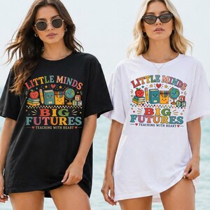 Peut inclure: Un t-shirt noir et un t-shirt blanc, chacun arborant un imprim&eacute; graphique color&eacute; avec le texte "LITTLE MINDS BIG FUTURES TEACHING WITH HEART" et des illustrations d'une pomme, de livres, de crayons, d'un globe et de cubes alphabet.