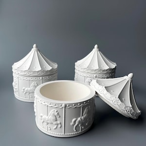 Op de afbeelding: Witte keramische sieradendoosjes met een carrouselthema. De dozen hebben een carrouselpaardenontwerp aan de zijkanten en een conisch deksel. Eén doos is open en onthult de binnenkant. De achtergrond is effen grijs.