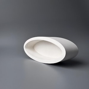 May include: A white, oval-shaped ceramic container with a smooth, matte finish. The vessel has a rounded, raised rim and a recessed interior. The object is set against a gray background, creating a minimalist aesthetic.