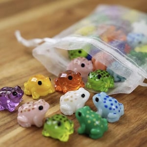 Peut inclure: Une collection de figurines de grenouilles miniatures colorées, de différentes couleurs, dont le violet, le jaune, l'orange, le vert, le blanc et le bleu. Certaines grenouilles sont éparpillées sur une surface en bois, d'autres dans un petit sac en filet blanc.