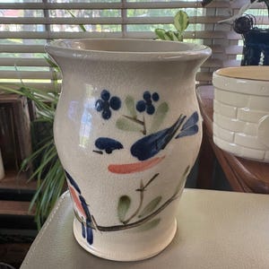 May include: A cream-colored ceramic vase with a crackle glaze finish, featuring hand-painted blue birds, green leaves, and small blue flowers on branches. The vase has a wide, flared rim and a tapered base, showcasing a decorative floral motif.