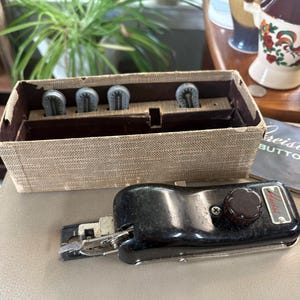 Può includere: Un accessorio per asole Greist nero con una manopola di regolazione marrone e componenti in metallo, accompagnato da una scatola rettangolare rivestita in tessuto beige contenente quattro camme in plastica grigia.