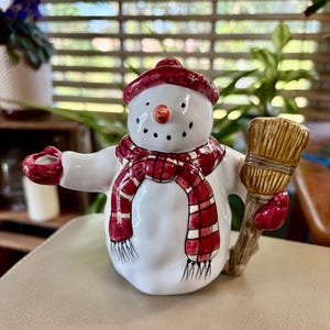 Peut inclure: Une figurine de bonhomme de neige en céramique blanche portant un bonnet rouge texturé et une écharpe à carreaux rouges et blancs. Le bonhomme de neige a un petit nez en carotte orange, des yeux et une bouche noirs, et tient un balai marron.
