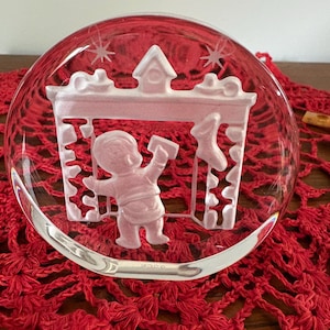 Puede incluir: Un adorno navideño de cristal transparente con un diseño esmerilado de un niño colgando una media. El adorno es redondo y se asienta sobre una superficie roja de ganchillo. El diseño incluye una chimenea y estrellas.