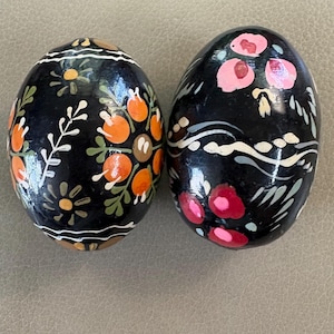 May include: Two oval-shaped decorative eggs with a glossy black finish. The left egg features hand-painted orange circular motifs and small yellow flowers. The right egg displays pink and red floral patterns with white wavy line accents.