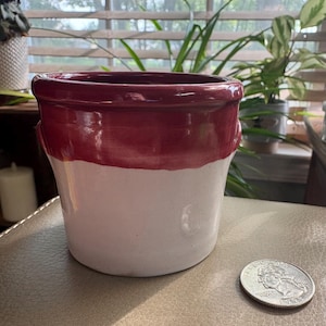 May include: A ceramic pot with a glossy deep red upper section and a matte off-white lower section. The pot features a wide, rounded rim and a slightly tapered cylindrical body. A silver-colored United States quarter coin is placed beside it.