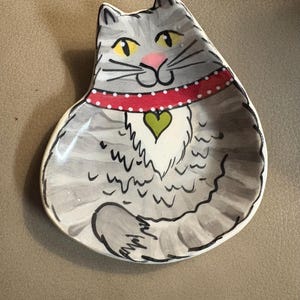 Puede incluir: Un plato de cerámica con forma de gato con pelaje gris a rayas, ojos amarillos y nariz rosa. El gato lleva un collar rojo con puntos blancos y un colgante verde en forma de corazón. El plato tiene un acabado brillante y contornos negros.