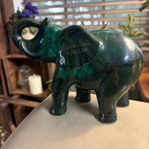 May include: A glossy dark green ceramic elephant figurine with its trunk raised upward. The side of the elephant's ear features gold cursive text that reads Niagara Falls Canada. The surface of the ceramic has a mottled, deep emerald finish.