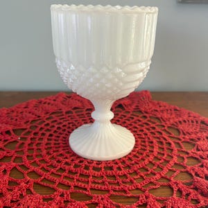 Puede incluir: Un cáliz de vidrio blanco de estilo vintage, con una base texturizada con patrón de diamantes y una sección superior acanalada. El cáliz se encuentra sobre una base de pedestal y sobre un tapete rojo tejido.