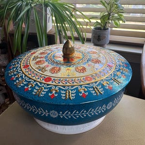 May include: A round, teal-coloured metal tin with a removable lid featuring a central gold-toned finial. The lid and sides are decorated with intricate, multi-coloured floral and scroll patterns in shades of red, white, and gold, resting on a white base.