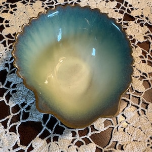 May include: A ceramic bowl shaped like a seashell with a scalloped edge. The interior features a gradient color transition, starting with a deep teal blue at the rim that fades into a soft cream color toward the center of the bowl's base.
