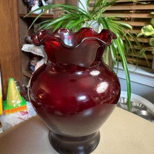May include: A deep red glass vase featuring a bulbous body and a wide, ruffled, crimped rim. The vase has a smooth, glossy finish and a circular base, standing upright with a consistent dark ruby color throughout the glass structure.