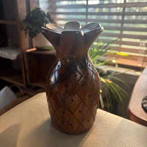 May include: A handcrafted wooden pineapple sculpture with a detailed, textured surface. The brown sculpture features a diamond pattern and small circular cutouts. The pineapple's crown is made of carved wooden leaves. The sculpture is displayed on a neutral-toned surface.