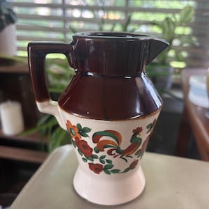 Puede incluir: Una jarra de cerámica con una sección superior de color marrón oscuro brillante y una base de color crema. La parte crema está decorada con un patrón pintado de colores que muestra un gallo, hojas verdes y flores naranjas y rojas.