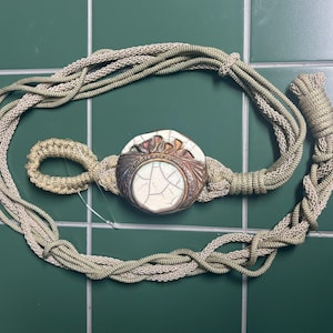 May include: A decorative curtain tieback featuring a braided light beige cord and a central circular ornament. The ornament has a white crackle-glazed center surrounded by a metallic bronze-toned border with sculpted leaf-like details.
