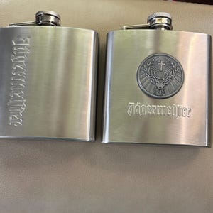 May include: Two brushed stainless steel liquor flasks with screw-top caps. The left flask features the vertical text Jägermeister. The right flask displays a circular emblem of a stag with a cross between its antlers above the text Jägermeister.